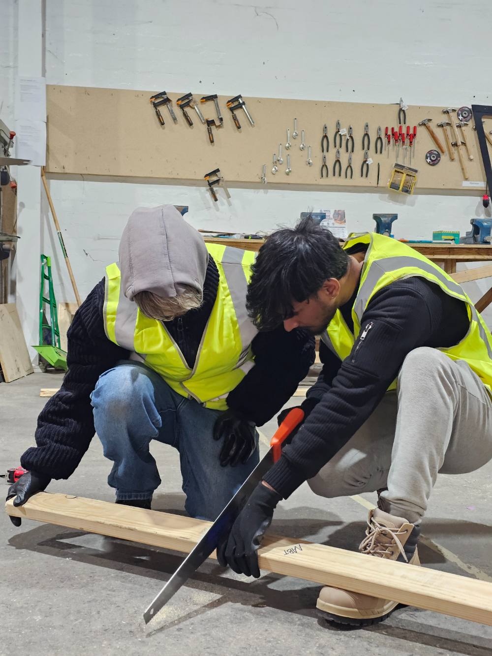 Two students cutting a length of timber with a handsaw on the workshop floor