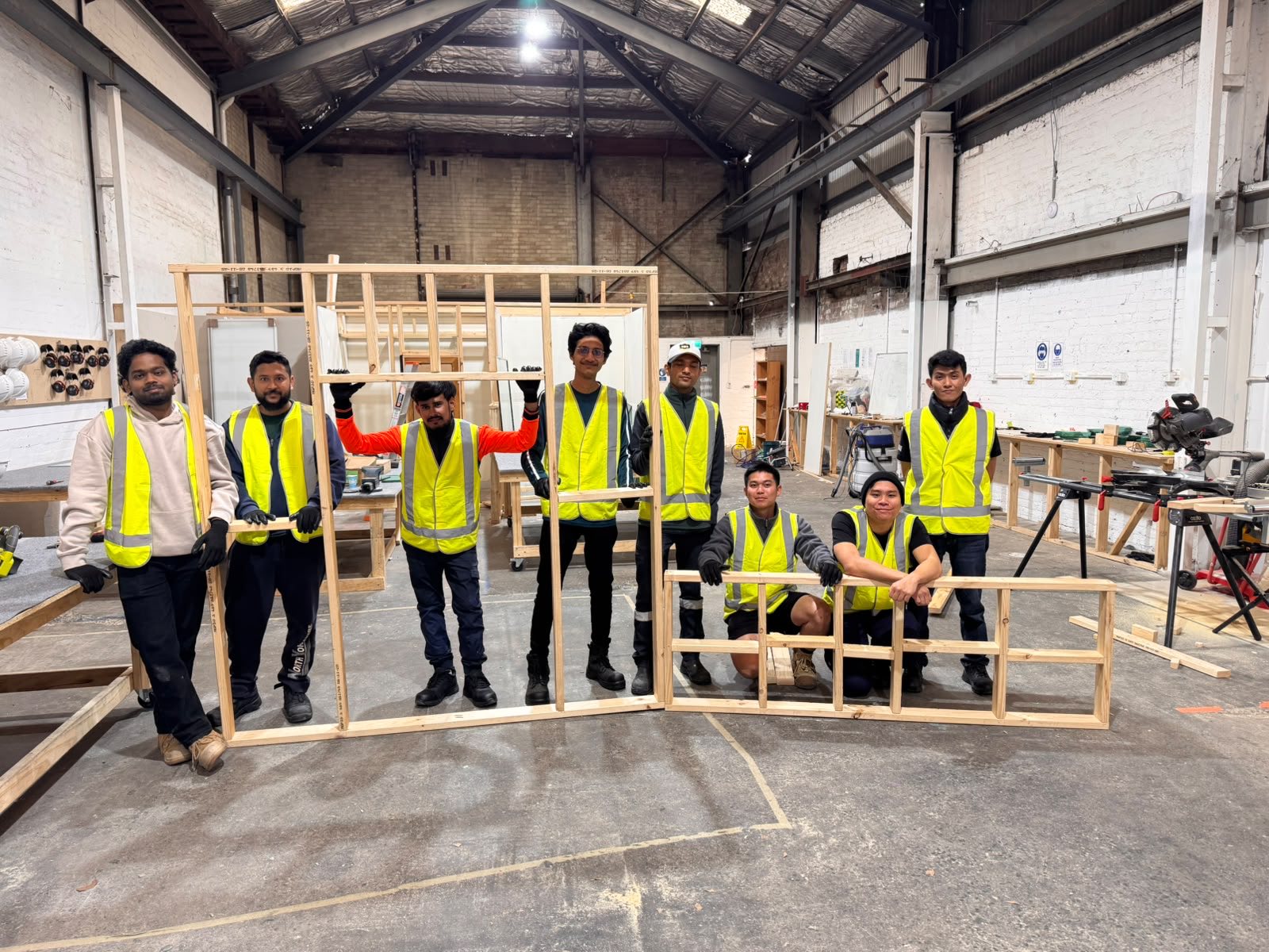 A group of construction students in high-visibility vests with the timber wall frames they built in the workshop