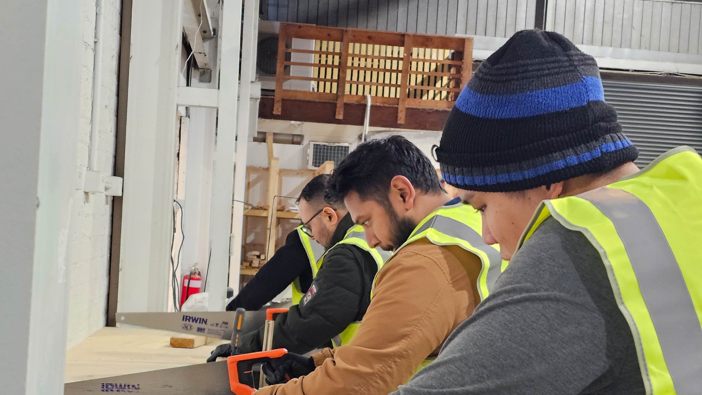 Students in high-visibility vests cutting timber with handsaws at a workbench in a training workshop