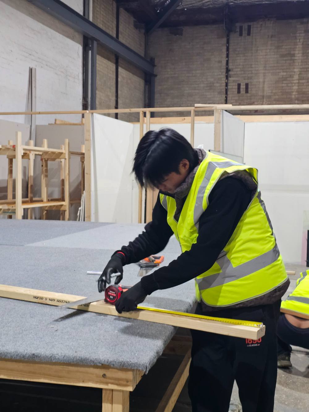 A student in a high-visibility vest measuring a length of timber with a tape measure
